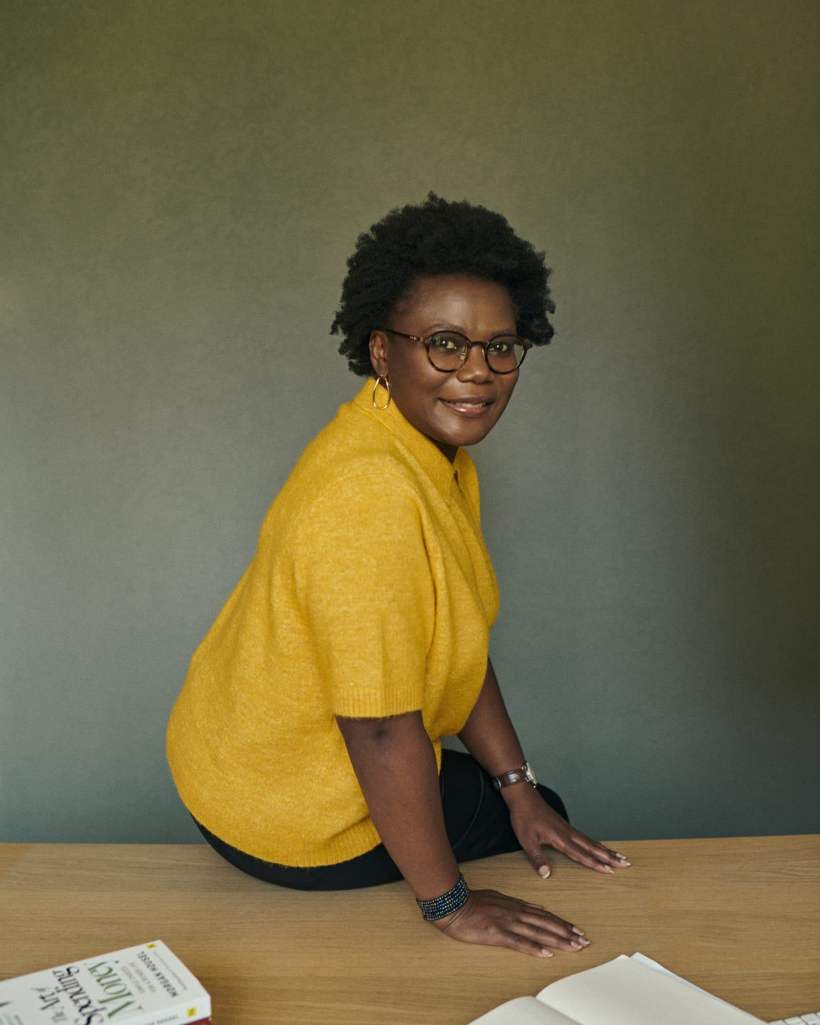 Alberta Opoku in een gele trui, zittend op een tafel met boeken over geld